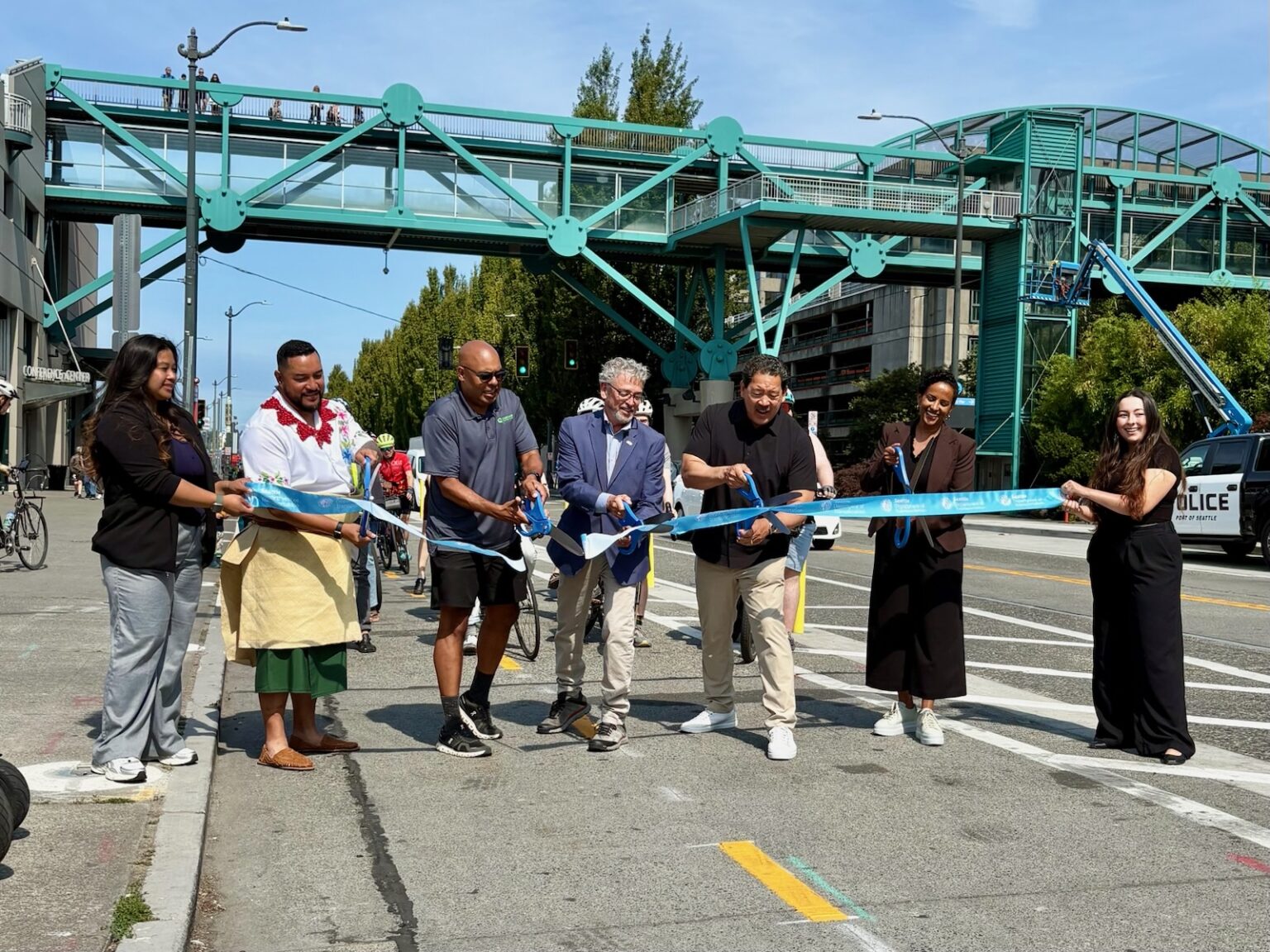 Several people cut a large ribbon at an outdoor celebration event on a sunny day, with a bike lane in the foreground.