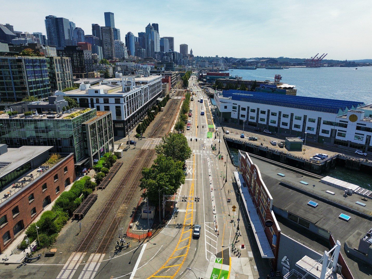 An aerial photo of a long street in the city, with train tracks to the left and buildings to the upper left. Water is shown to the upper right.