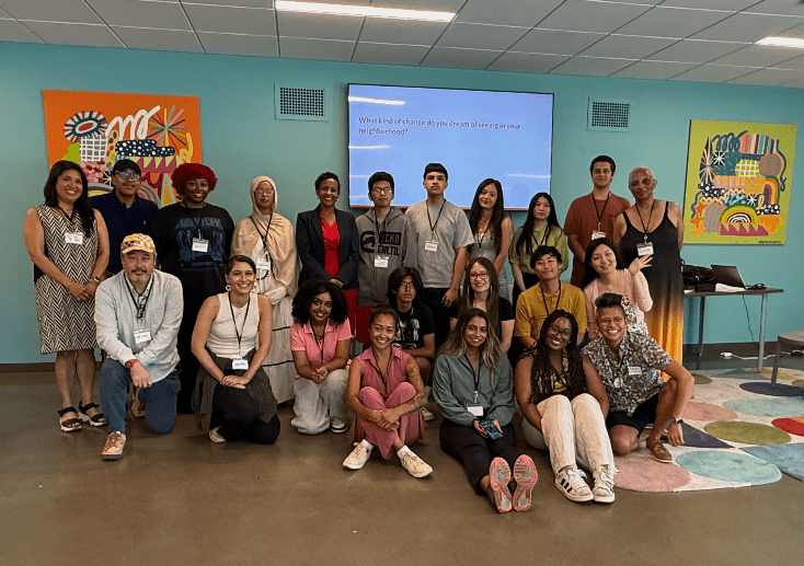 A large group of people in an indoor meeting space smile at the camera. A large screen is in the background.