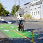 A person wearing a helmet and white t-shirt bikes on a bike lane in the city with buildings and trees in the background. The bike lane is marked in green paint where it travels past a driveway.