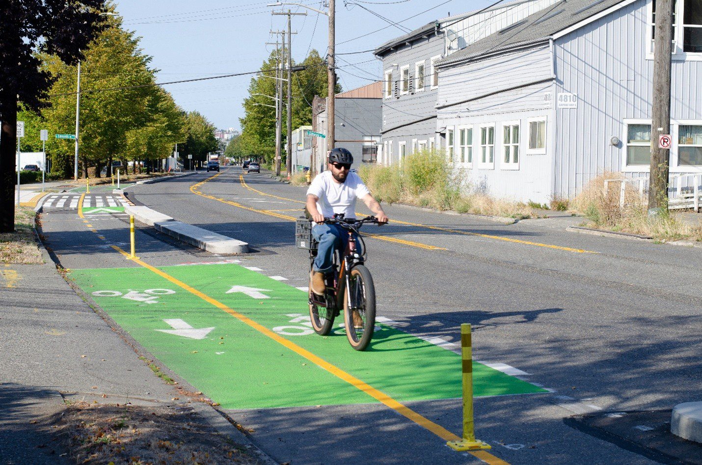 A person wearing a helmet and white t-shirt bikes on a bike lane in the city with buildings and trees in the background. The bike lane is marked in green paint where it travels past a driveway.