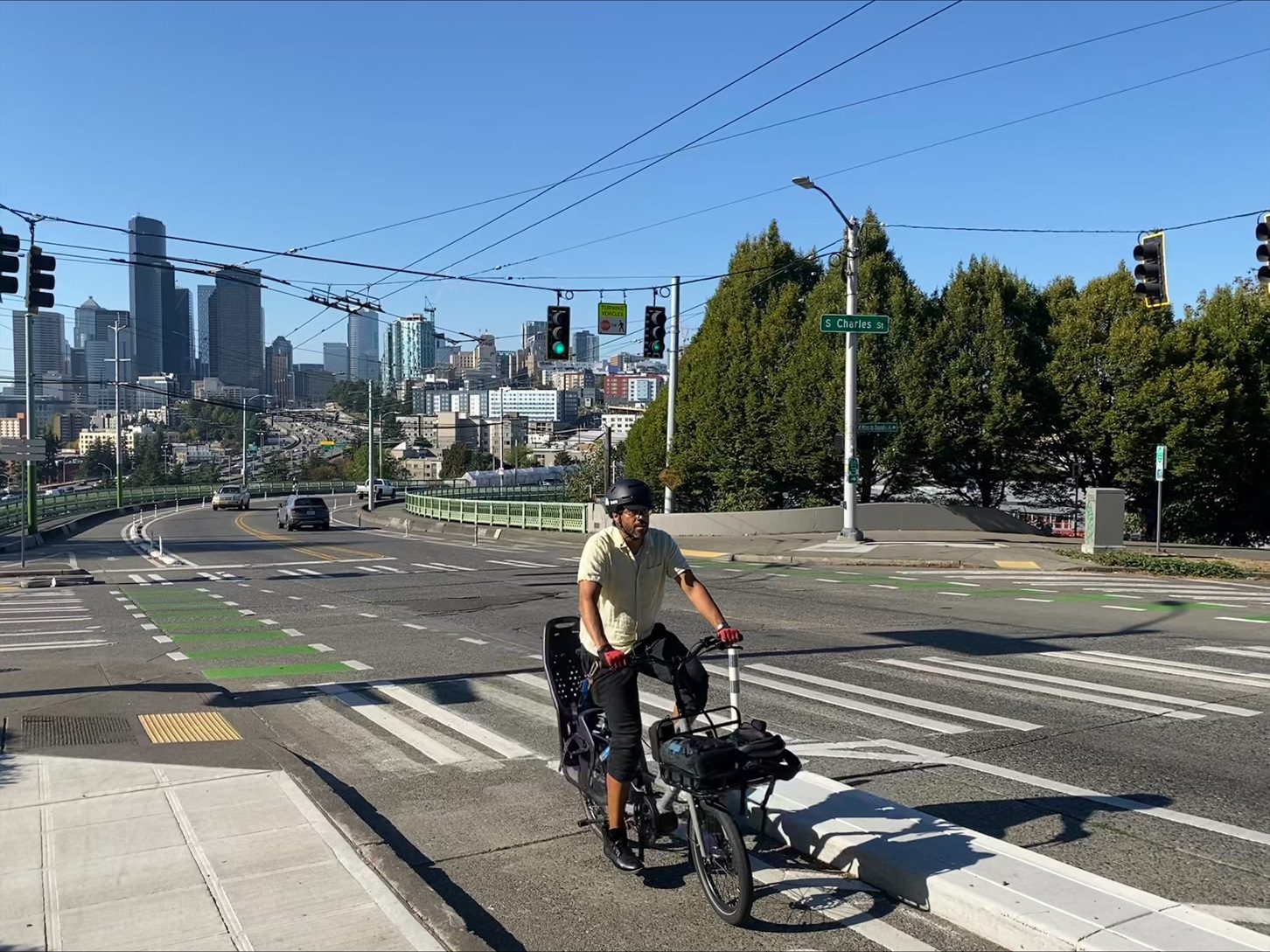 A person rides a bike up a hill in a protected bike lane with a cityscape and trees in the background.