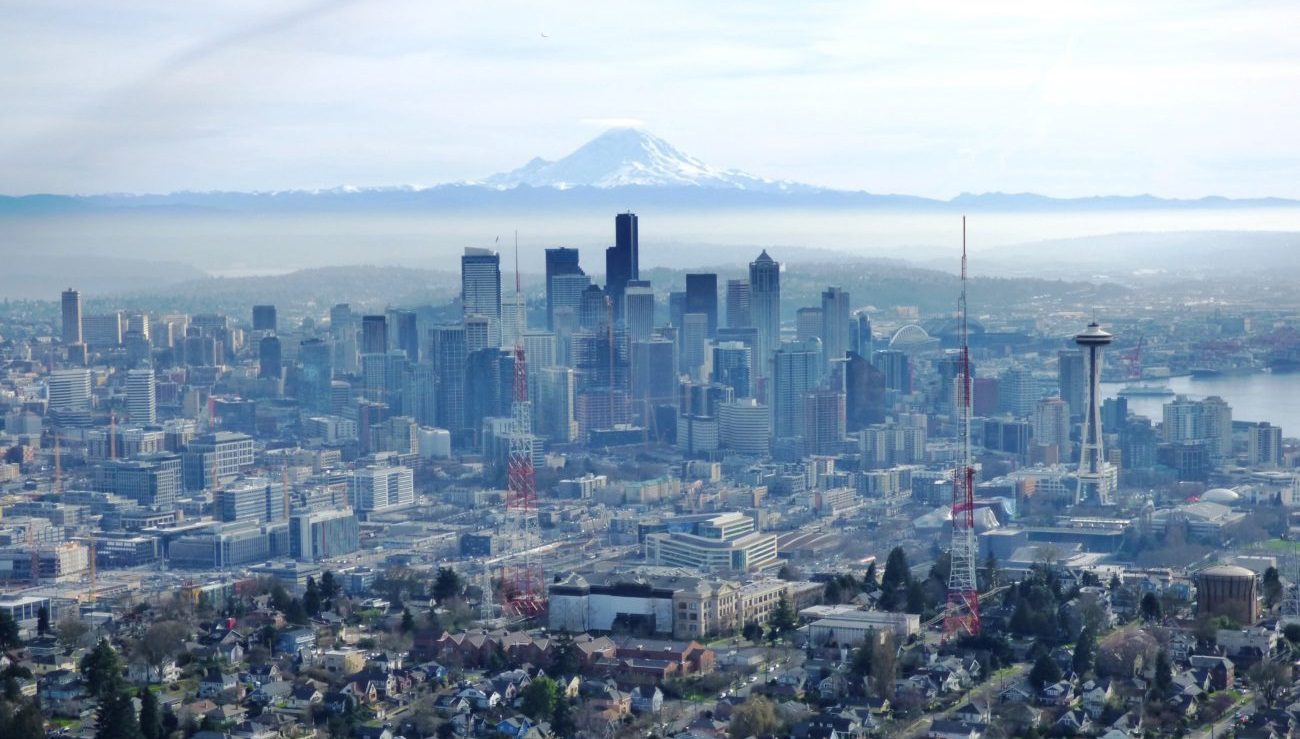 An aerial photo of the city of Seattle looking towards downtown towers, with Mount Rainier in the background on a mostly cloudy day.