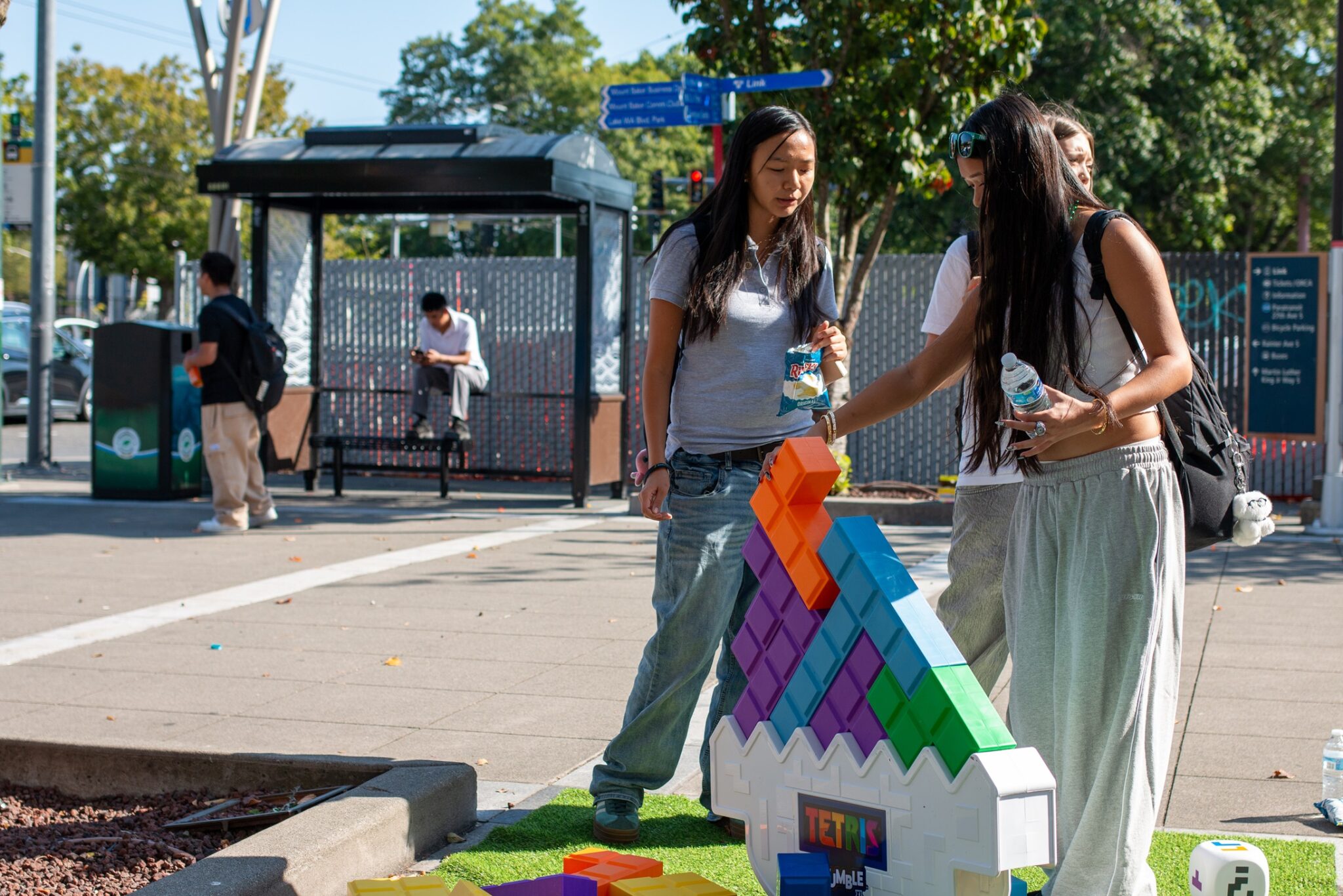 High school students play with a large game of Tetris blocks while standing outside on a sunny day. People wait for a bus in the background.