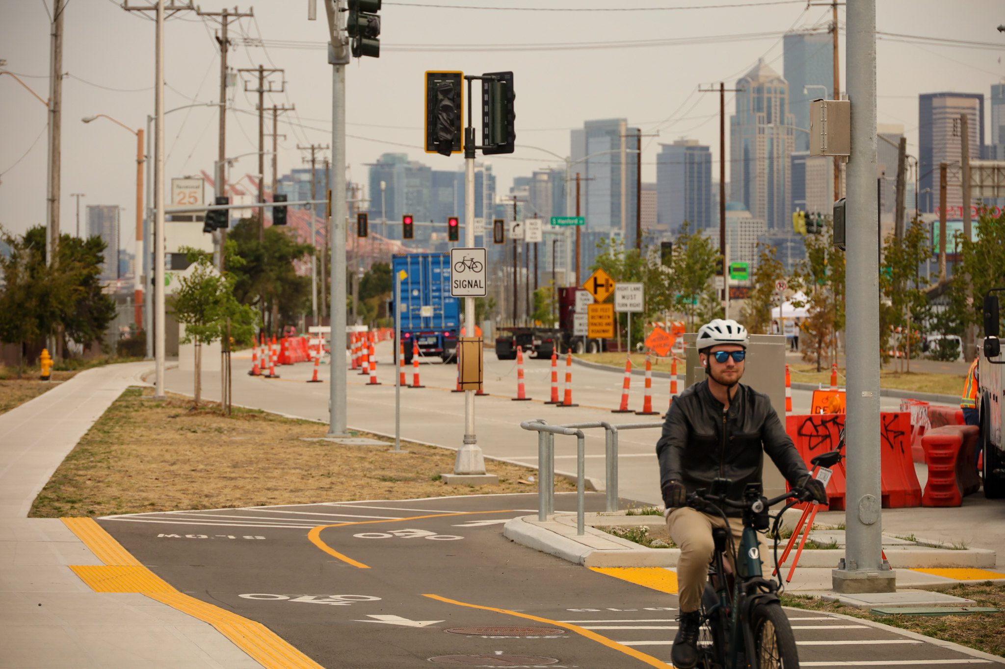 A person wearing a white helmet bikes towards the camera on a section of protected bike lane. In the background a freight truck, signs, cones, and large skyscraper buildings are visible.