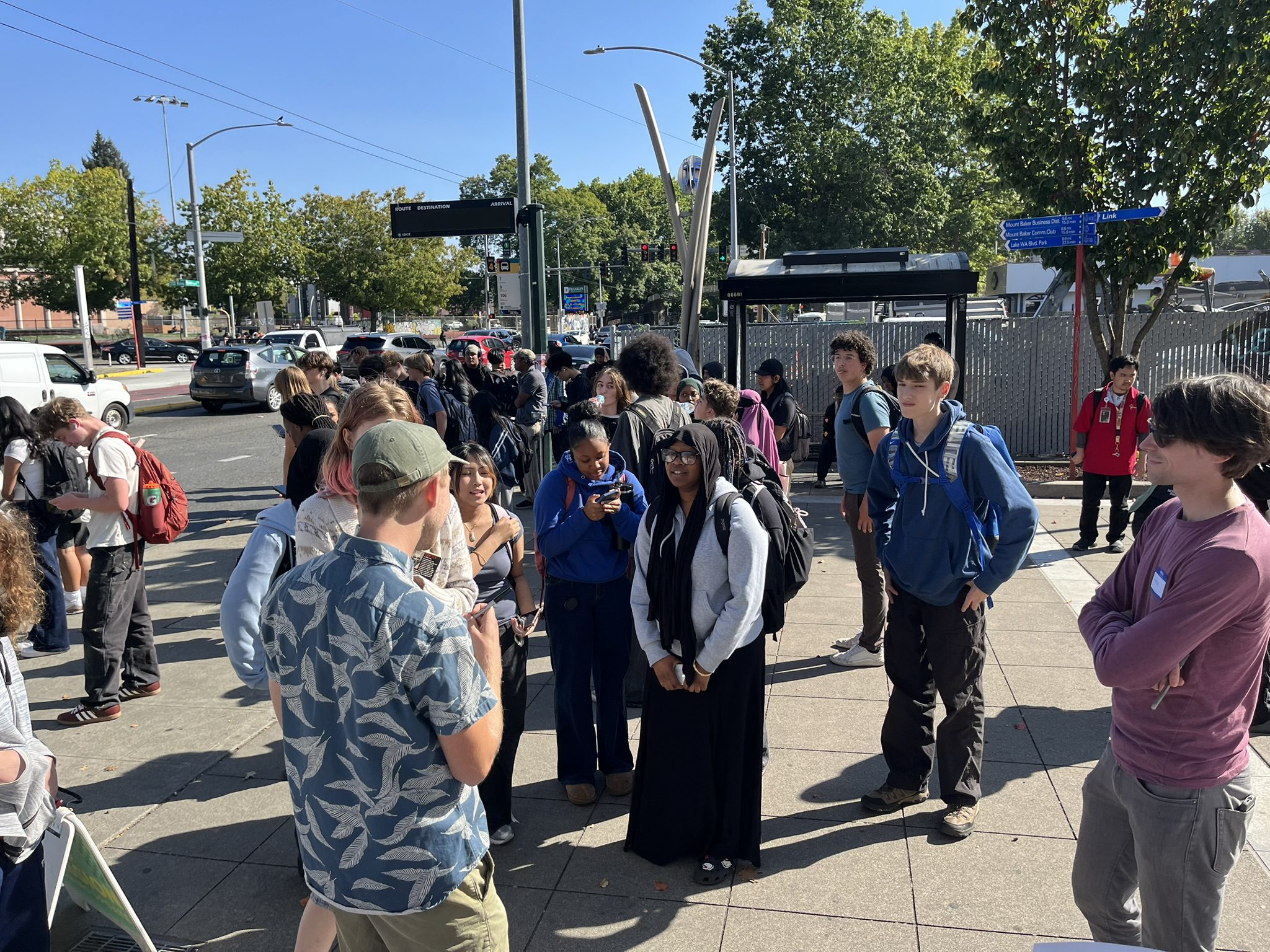 A large crowd of people standing on a plaza area on a sunny day. Many young people are waiting for the bus. Large trees and blue skies are in the background.