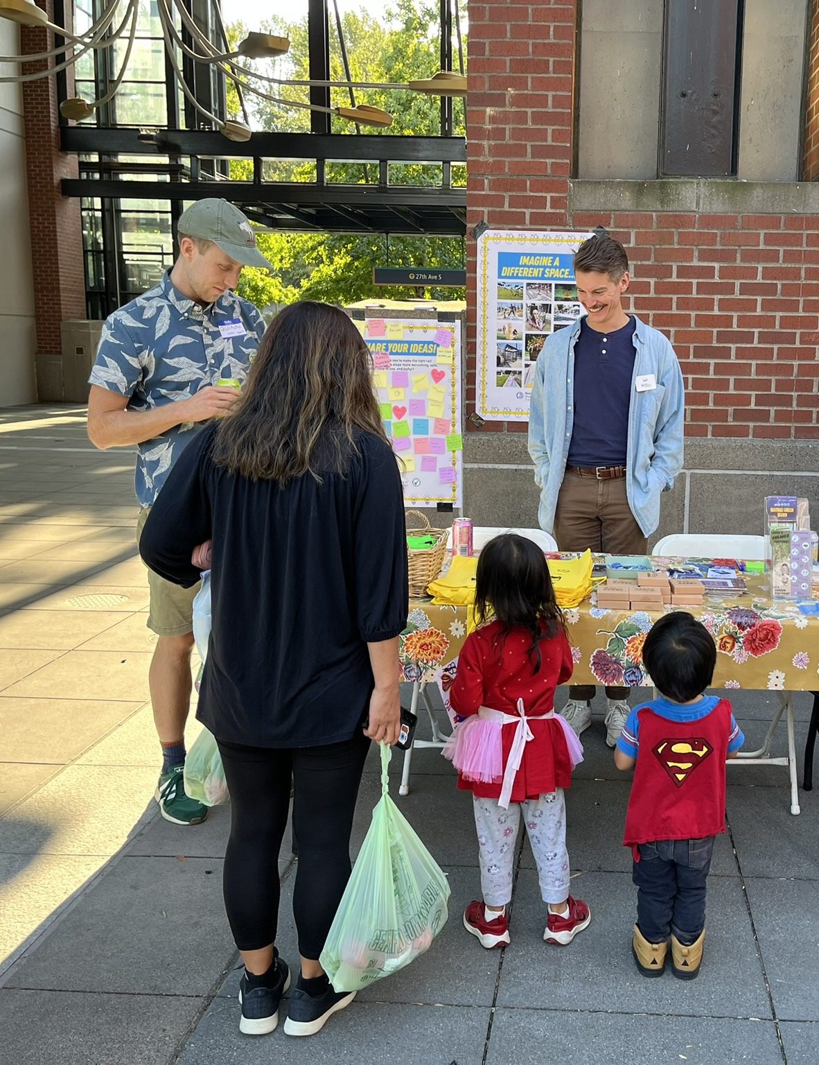 Two people speak with an adult and two kids at an outdoor event, in front of a table with materials and signs in the background.