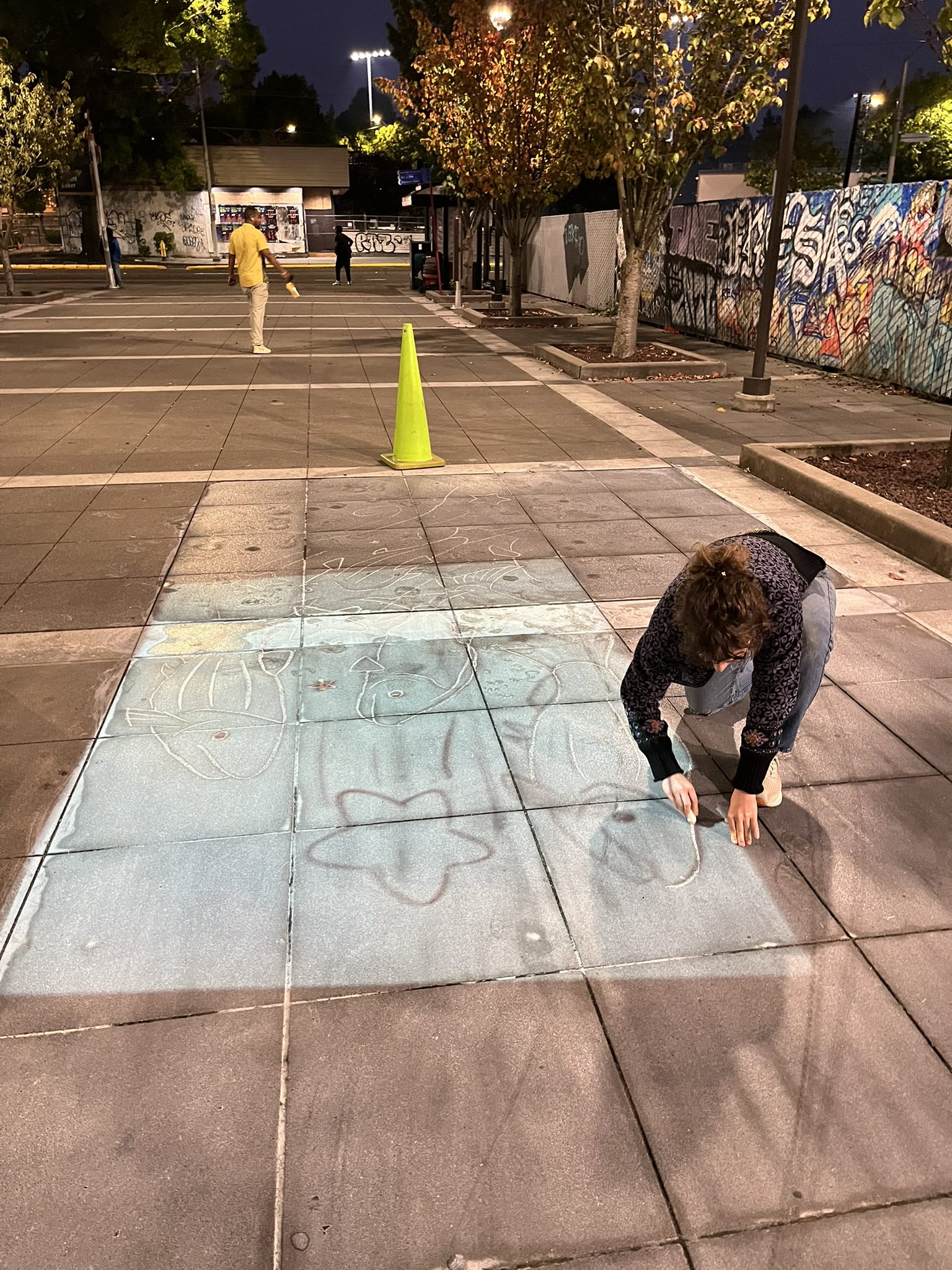 A person paints in the outlines of an artwork on a large open plaza space.