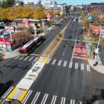 An aerial view of a city street with crosswalks, bus lanes, a bus, trees, buildings, a center median, traffic signals, sidewalks, and other city elements in the picture.