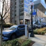 Several parked cars are alongside a curb space in the city. Trees, vegetation, large buildings and other city elements are in the image. A blue and white sign says "Pay 8am-8pm".