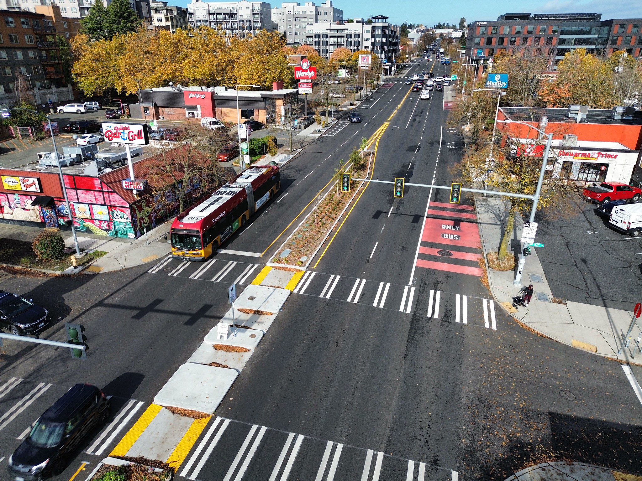 An aerial view of a city street with crosswalks, bus lanes, a bus, trees, buildings, a center median, traffic signals, sidewalks, and other city elements in the picture.