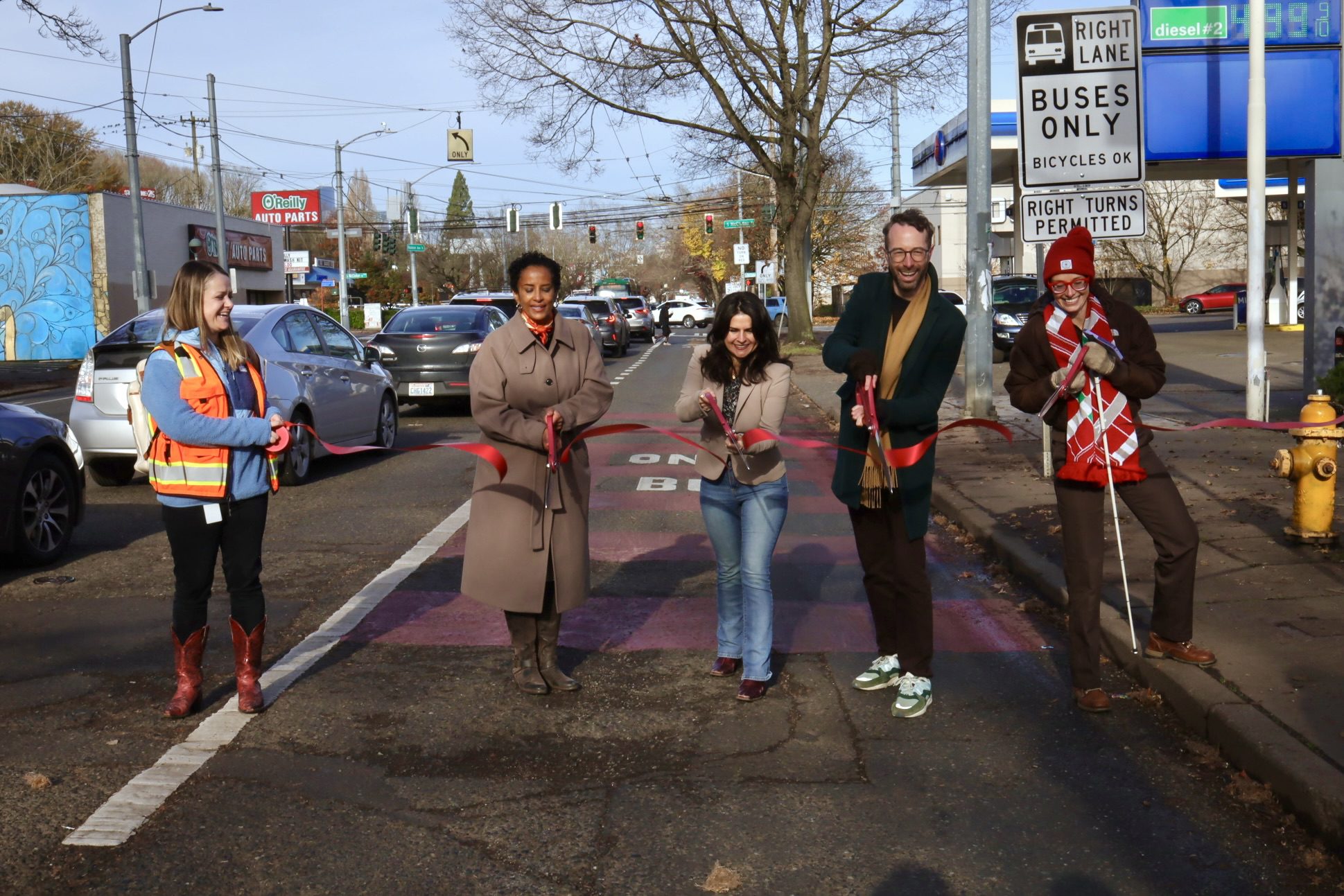 A group of several people cut a ribbon in a red bus lane on a clear, sunny day. A tree is above and several cars are to the left.