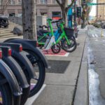 A row of parked rental bikes, including green Lime and blue Bird e-bikes, line a city sidewalk. A cyclist in a yellow jacket rides along a bike lane, surrounded by urban buildings.