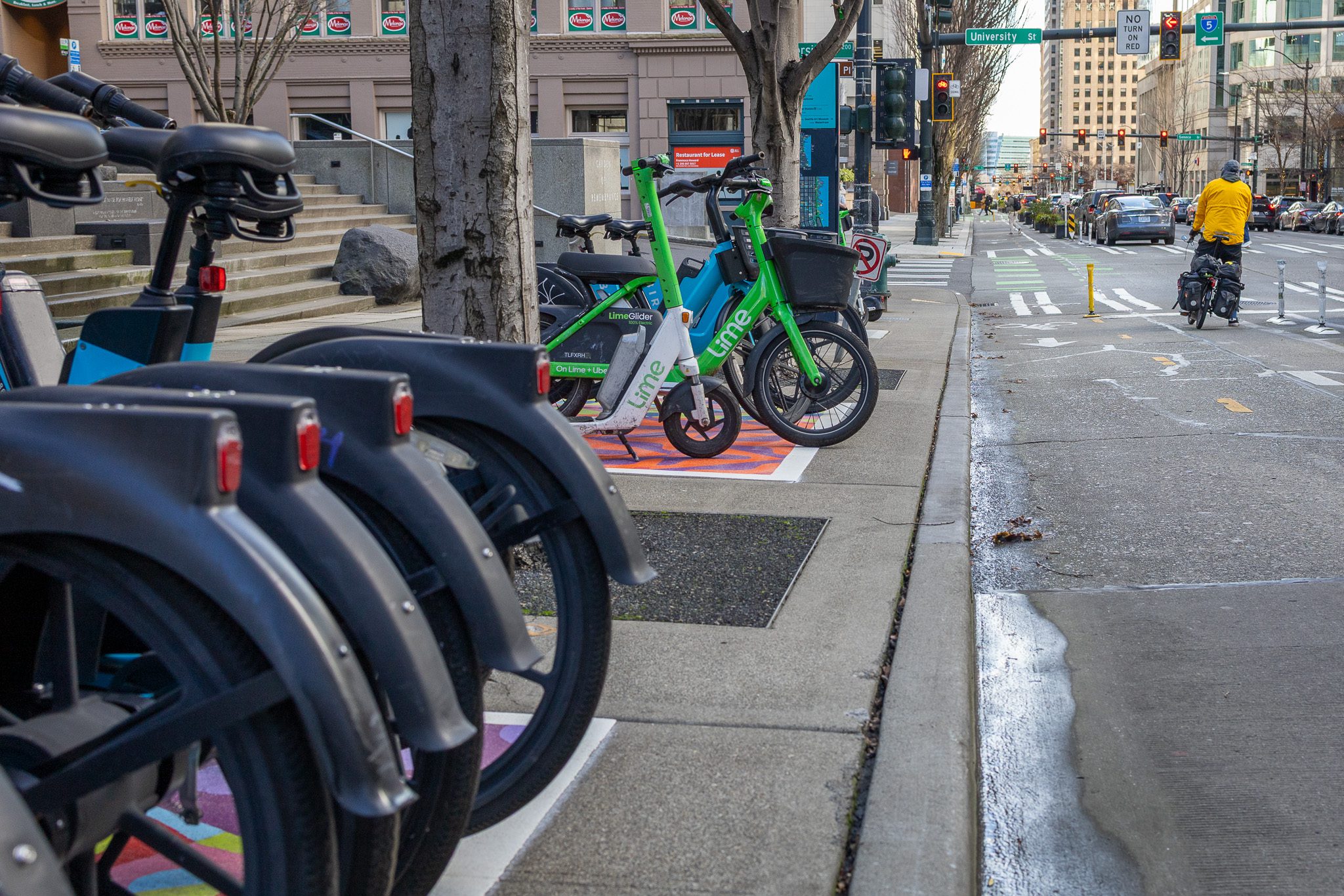 A row of parked rental bikes, including green Lime and blue Bird e-bikes, line a city sidewalk. A cyclist in a yellow jacket rides along a bike lane, surrounded by urban buildings.