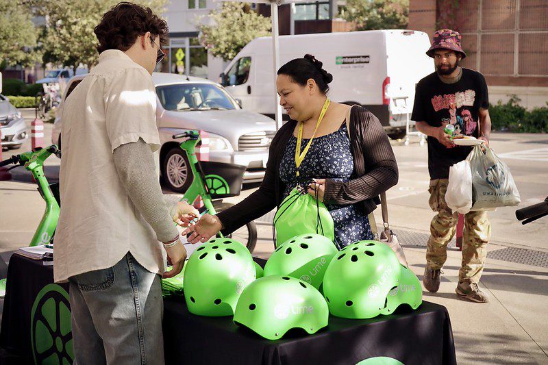 A woman and a man interact at a table with Lime scooters and bright green helmets. The scene is casual and outdoor.