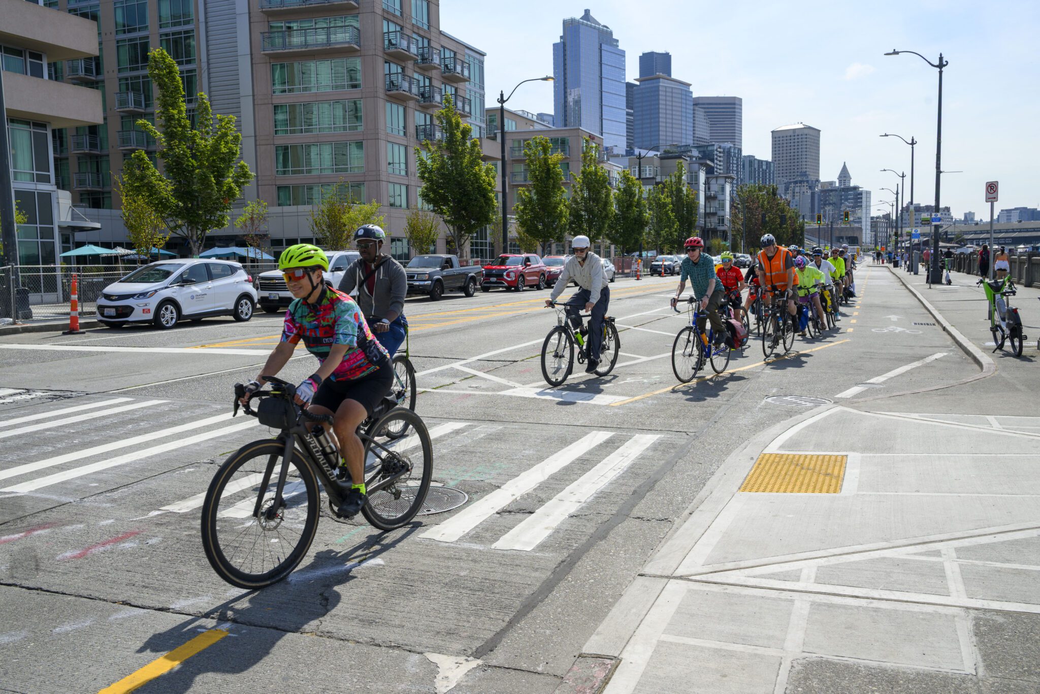 A group of cyclists using a marked bike lane on a city street. They're wearing helmets and colorful attire, with urban buildings and trees in the background. The mood is active and communal.
