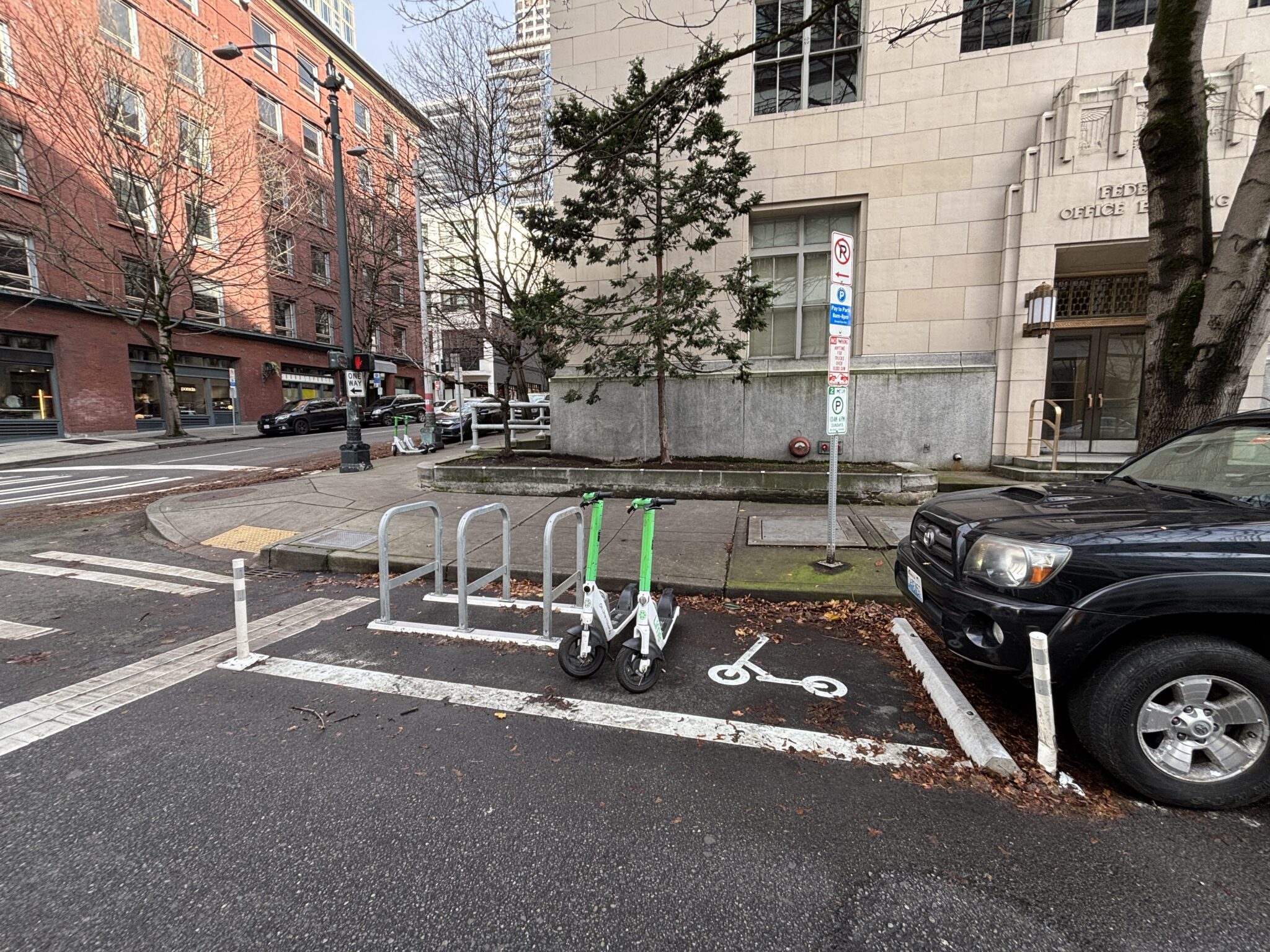 Two green and white scooters parked at a bike rack beside a no-parking sign on the street. An SUV is parked behind the scooter parking area, leaving a clearer space close to the sidewalk.
