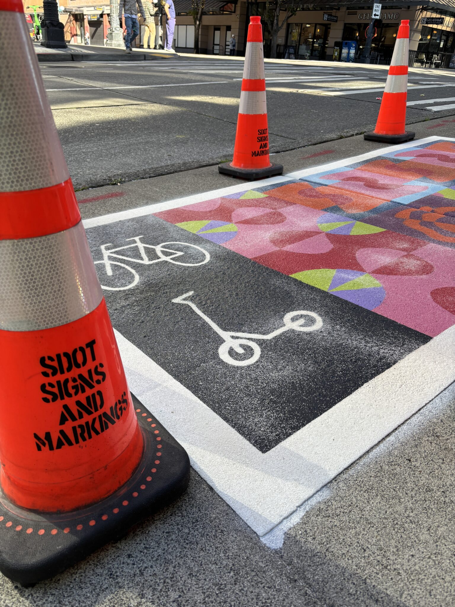 Orange cones line a colorful sidewalk with bike and scooter symbols, indicating a place to park shared devices. Pedestrians and traffic are visible in the background. A cone in the foreground has black text that says: "SDOT SIGN AND MARKINGS"