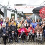 A large group of people sit and stand in front of a streetcar train that includes the words "Filipinotown" as well as artwork decals on the outside of the train.
