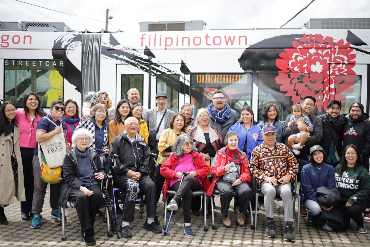 A large group of people sit and stand in front of a streetcar train that includes the words 