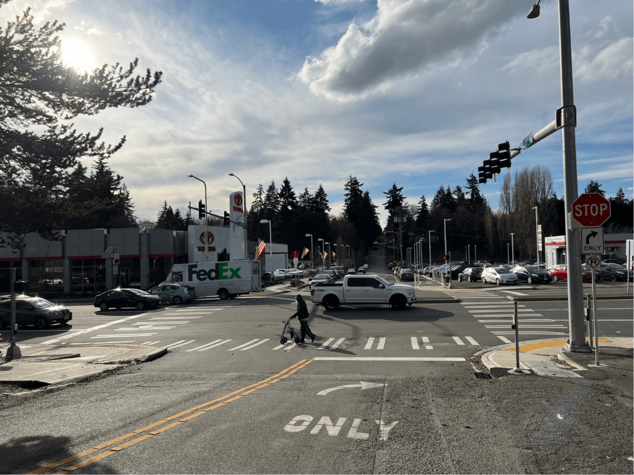 A person crosses the street at a marked crosswalk as several cars travel by on the street. Large trees and clouds are in the background.