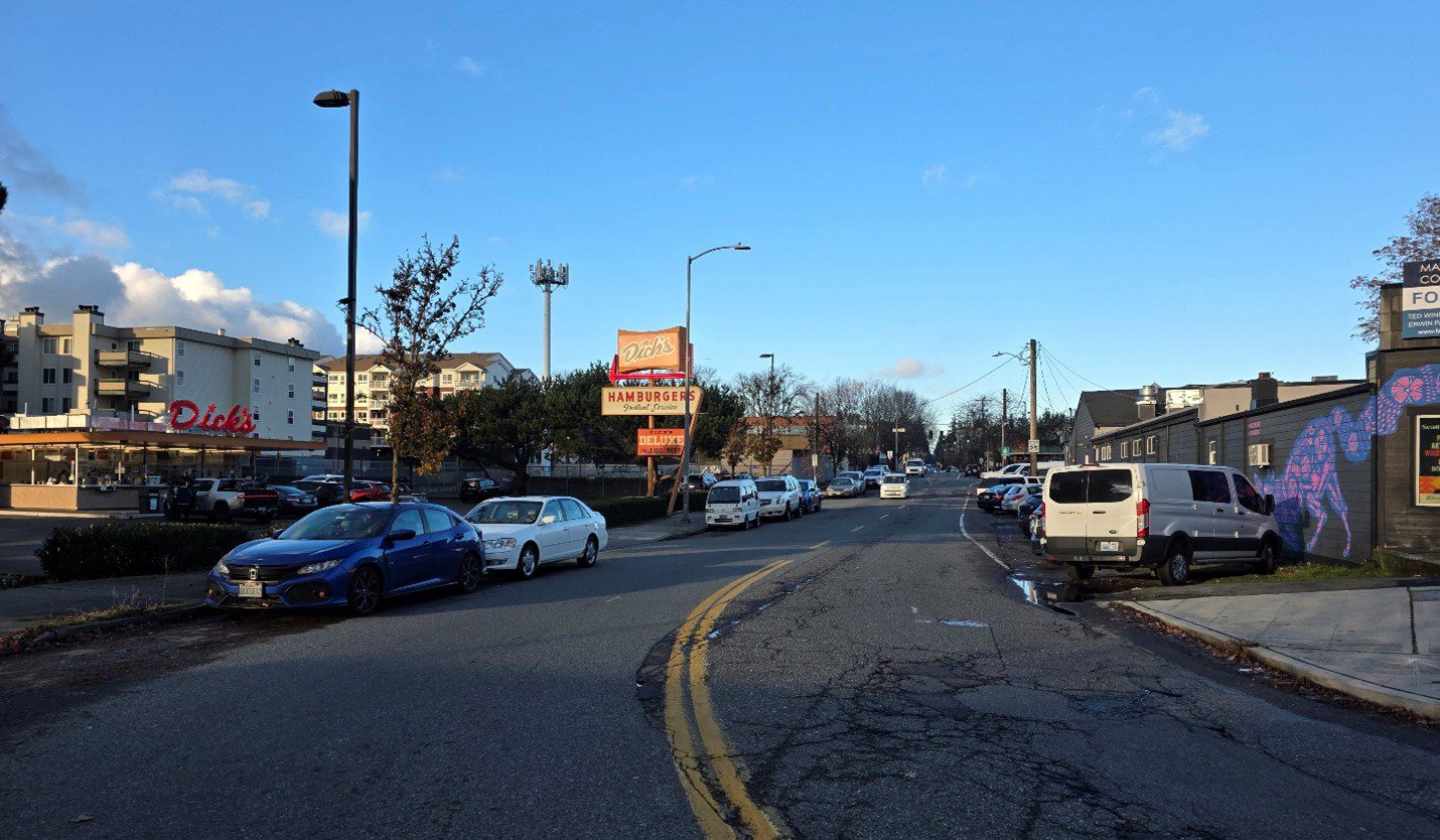 Photo of parked cars along a street on a mostly sunny day. Buildings and local businesses are in the background.