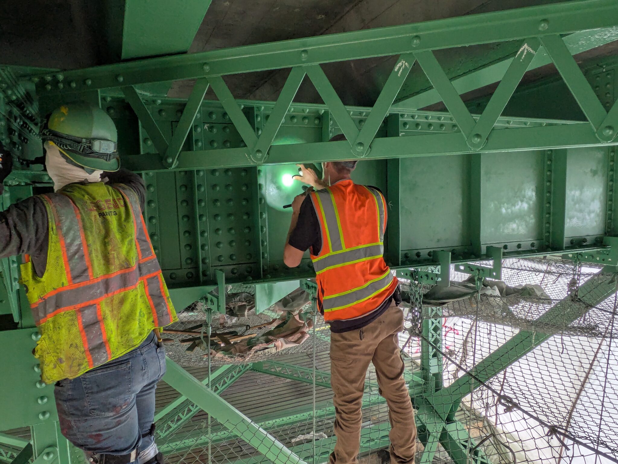 Two people work under a bridge. They are wearing safety vests. One person on the right has a flashlight shining onto the bridge structure.