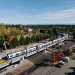Aerial image of a large light rail train with blue skies and trees in the background and a parking lot in the foreground.