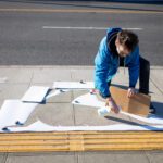 Man kneeling on a sidewalk holds a spray bottle in his right hand and a piece of cardboard in his left. He is looking down at the ground where there is a stencil cut in the shape of a seal.