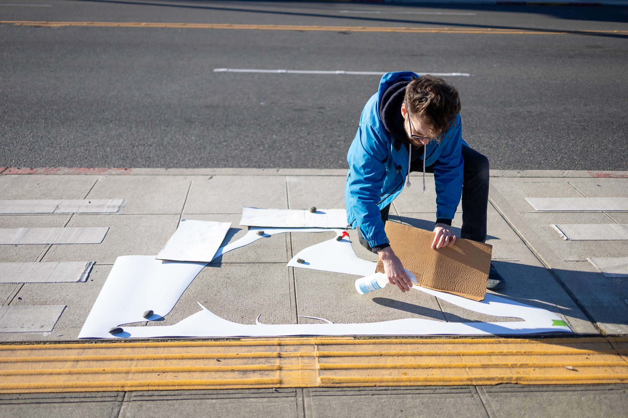 Man kneeling on a sidewalk holds a spray bottle in his right hand and a piece of cardboard in his left. He is looking down at the ground where there is a stencil cut in the shape of a seal.