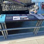 A photo of an interpretive sign with boats and water and a dock below.