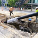 Construction workers install a large pipe into a trench in the street.