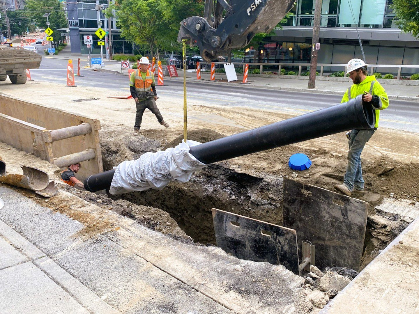 Construction workers install a large pipe into a trench in the street.