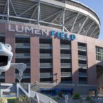A puppet salmon figure in front of a large stadium with the words Lumen Field on the front of the building, on a clear sunny day.