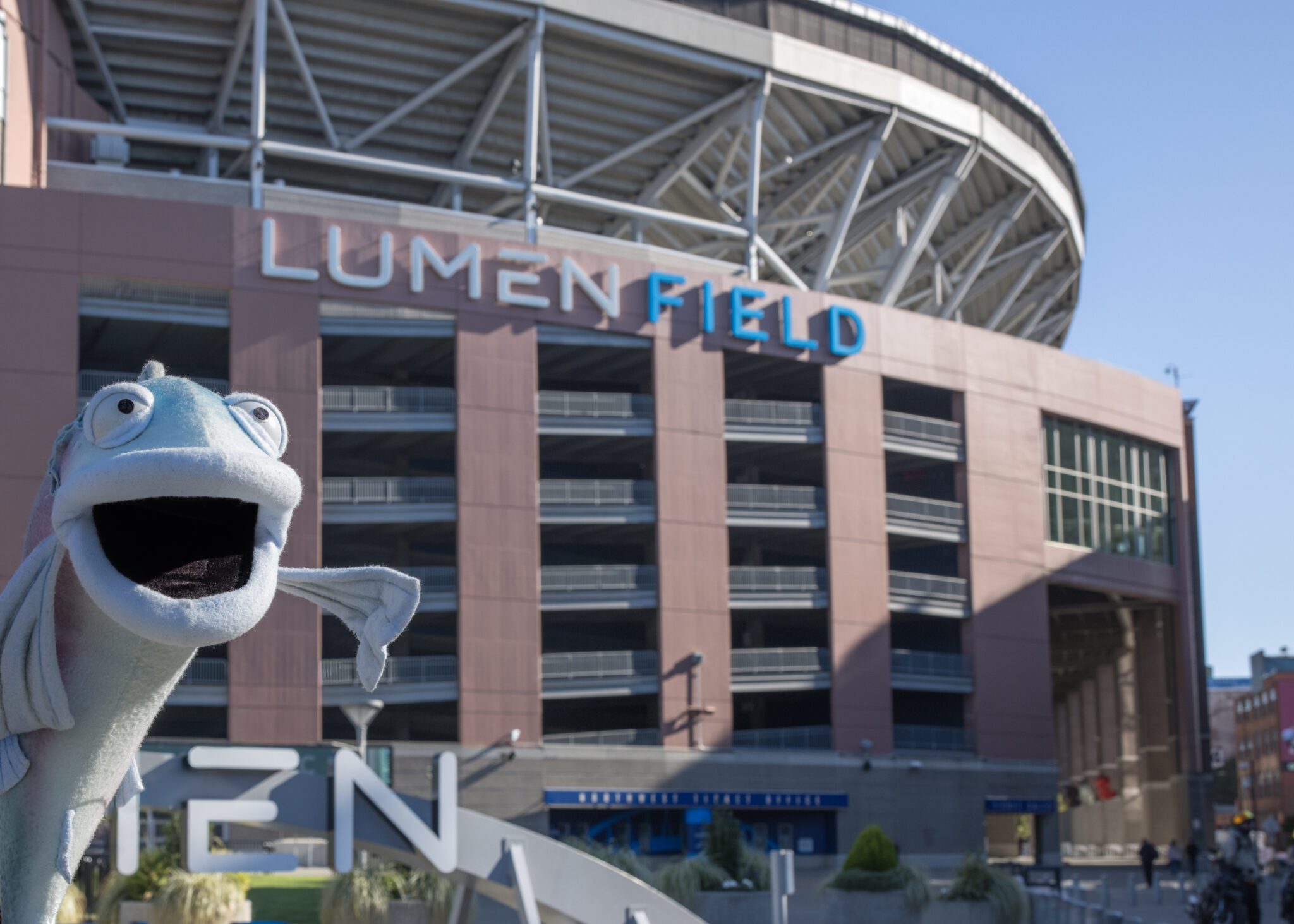 A puppet salmon figure in front of a large stadium with the words Lumen Field on the front of the building, on a clear sunny day.