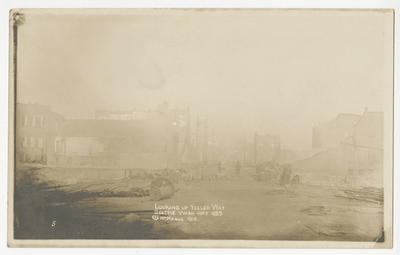 Sepia-toned photo from July 1889 showing the aftermath of the Great Seattle Fire on Yesler Way. The unpaved street is littered with debris, including wood planks, pipes, and a spoked wheel, while workers clear rubble and begin reconstruction. A partially intact building stands on the left, with damaged structures and haze obscuring the background.