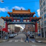 Ornate red and blue gate with traditional Chinese architecture spans a city street. A person crosses the road inside a crosswalk below, surrounded by urban buildings.