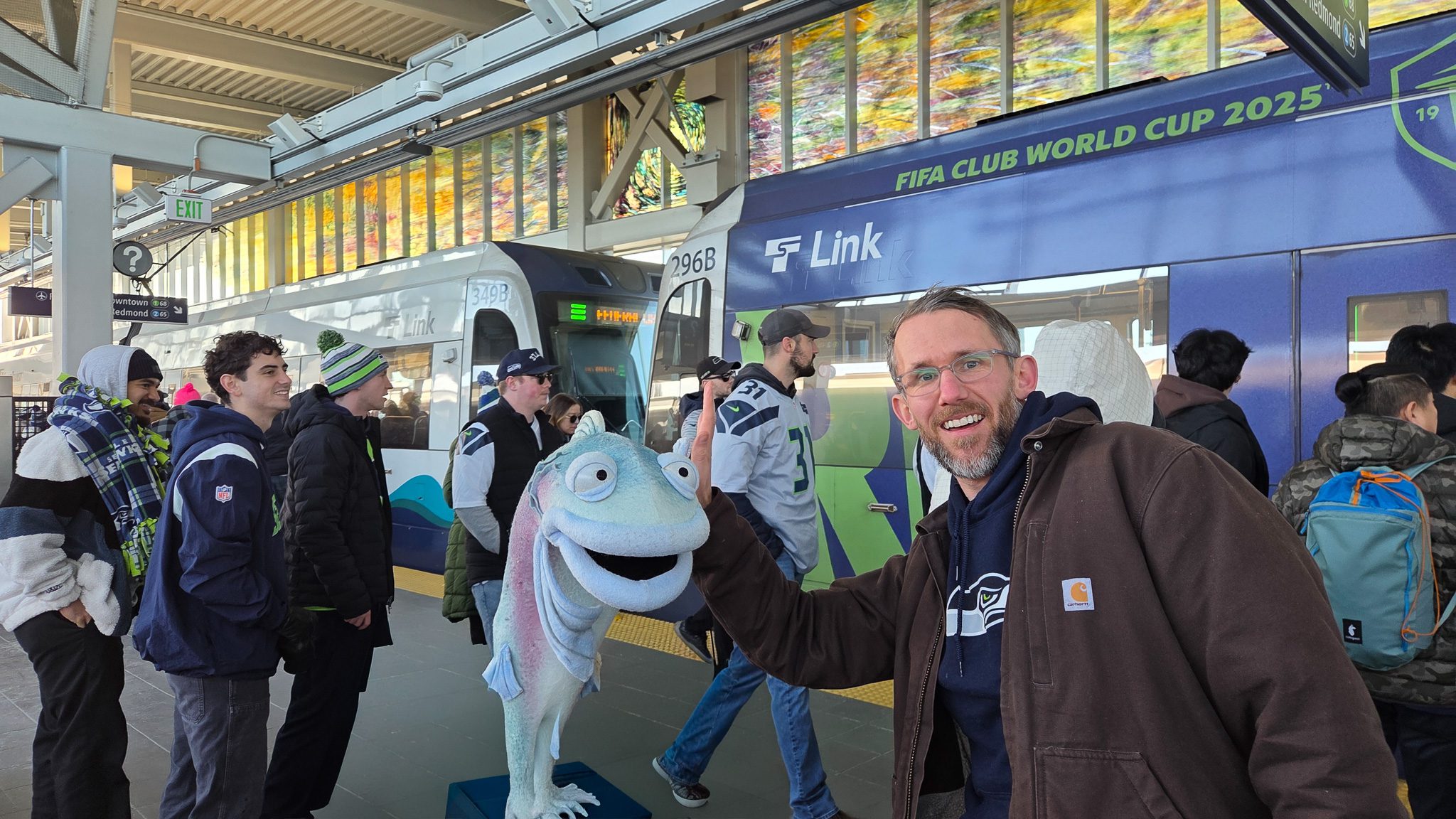 A salmon puppet and a man wearing a Seahawks sweater high five. Both are smiling at the camera. In the background, a crowd of people board a light rail, most of them are wearing Seahawks clothes.