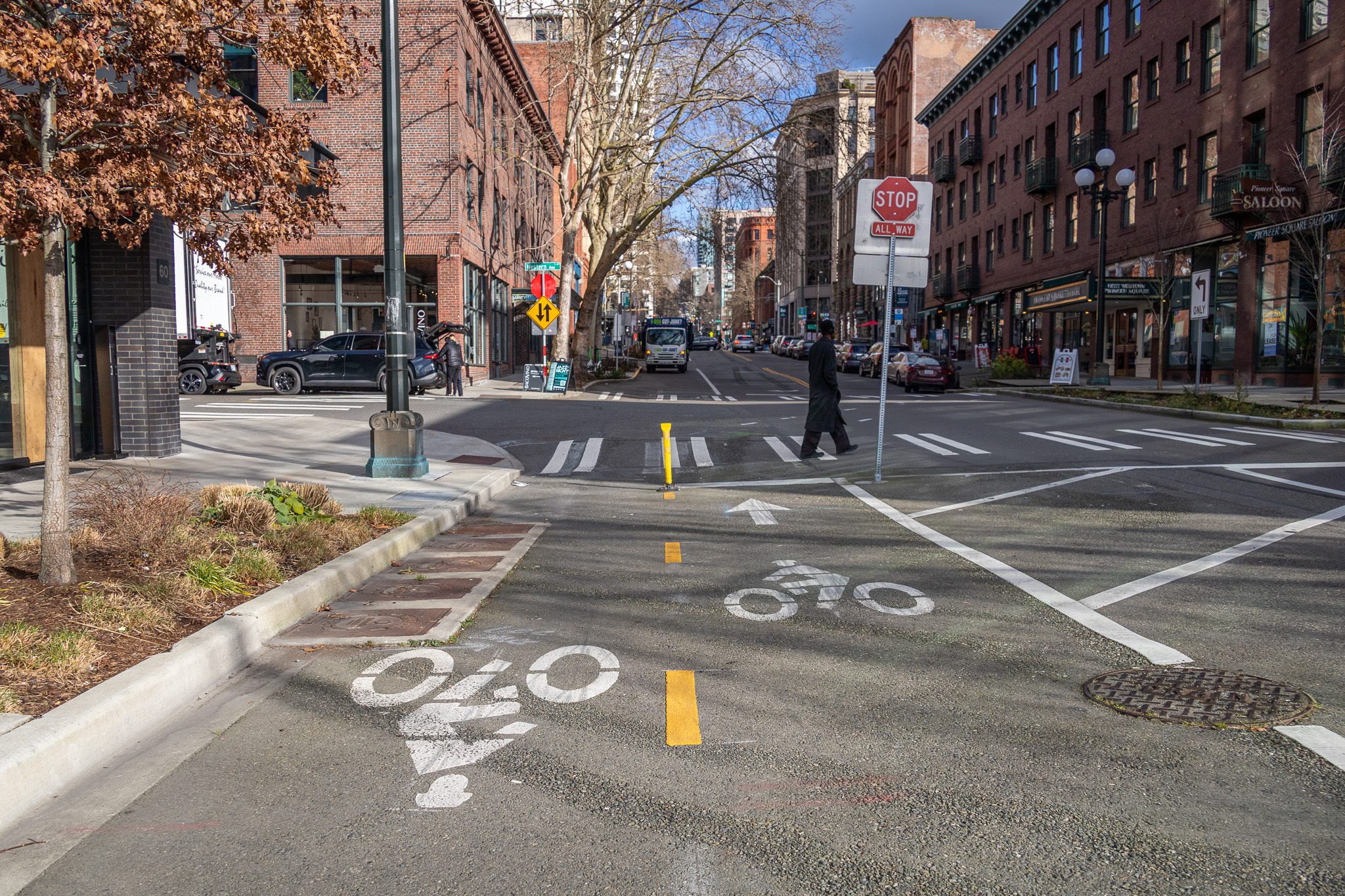 Street view of Pioneer Square with historic brick buildings, a bike lane, crosswalk, and a 'STOP ALL WAY' sign. A pedestrian crosses as vehicles line the street.