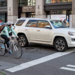 A busy urban street. A cyclist wearing a white helmet and teal shirt riding a light-colored road bike is stopped behind a crosswalk. She is next to a white SUV.