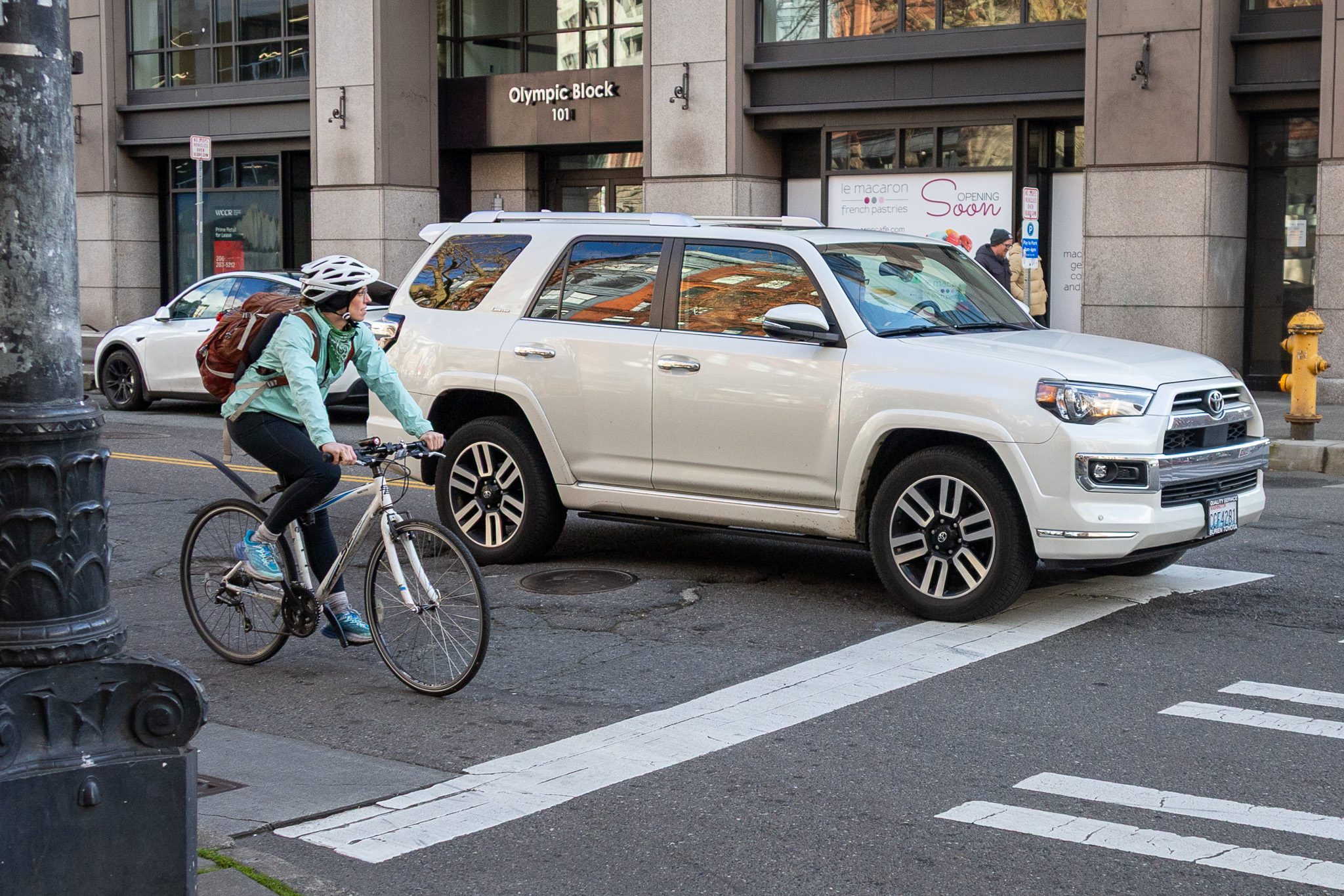 A busy urban street. A cyclist wearing a white helmet and teal shirt riding a light-colored road bike is stopped behind a crosswalk. She is next to a white SUV.