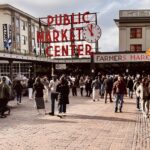 A bustling street scene at Pike Place Market in Seattle features the iconic red "Public Market Center" neon sign with a clock, the "Farmers Market" entrance, and a cobblestone street filled with people in winter attire. Buildings, street signs, and a lively crowd add to the vibrant atmosphere.