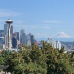 Panoramic view of Seattle's skyline on a sunny day, featuring the iconic Space Needle, modern skyscrapers, and Mount Rainier in the background, with lush green trees in the foreground.