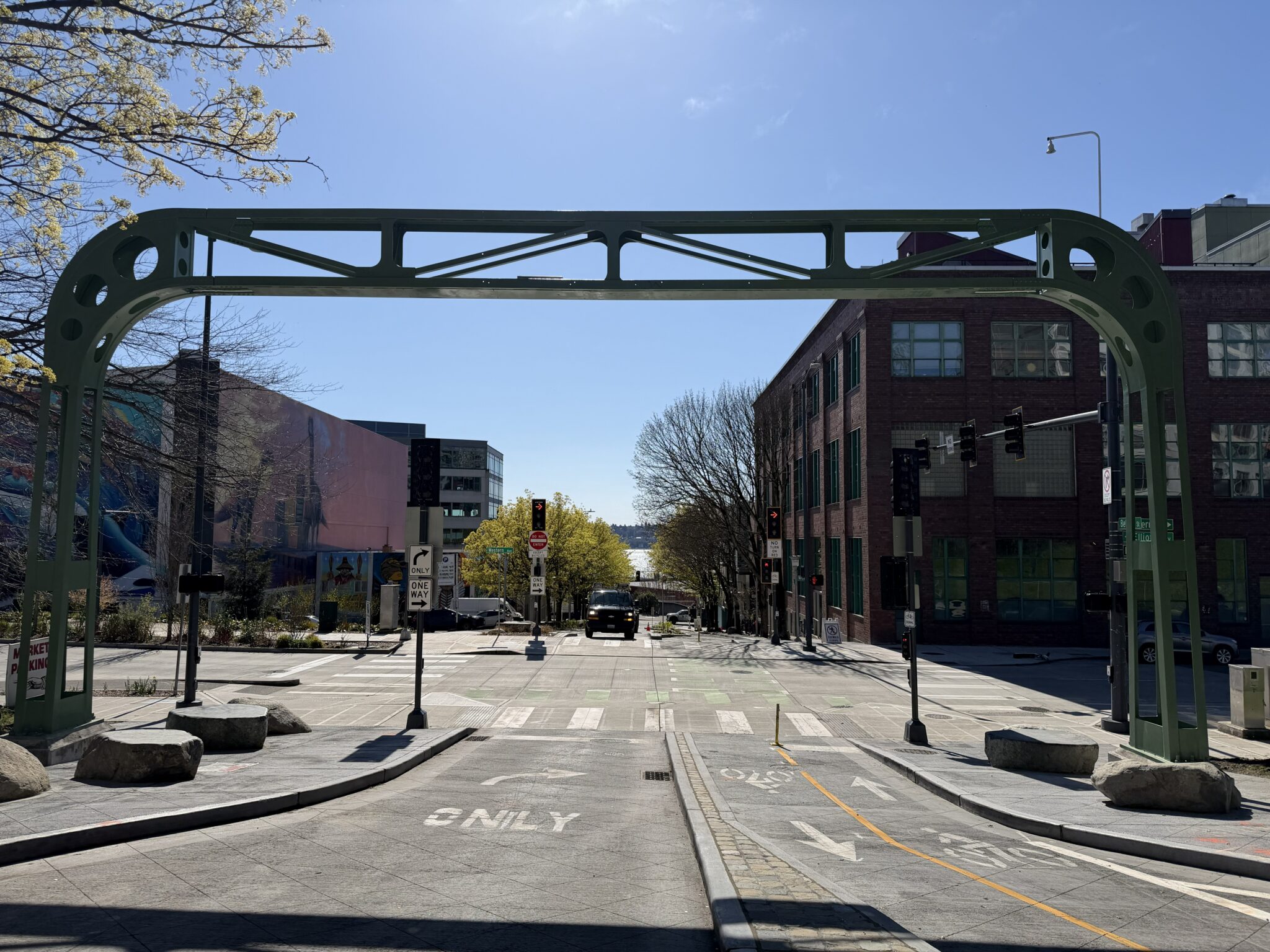 A road with a bike lane on the right and a vehicle lane with an arrow icon on the left. There is a median between the bike and auto lanes. A metallic arch spans the street and the adjacent sidewalk. Large flattop boulders sit at the base of the arch on either side.