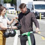 A person holding three bike helmets hands one to a man standing on a scooter inside a protected bike lane. A van and several cars are on the street in the background on the other side of the bike lane’s barrier.
