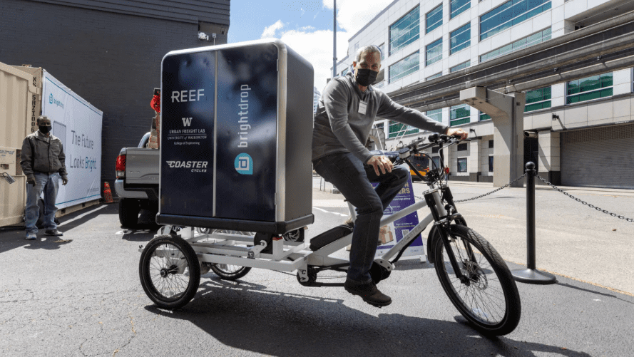 An outdoor scene featuring a man riding a white cargo e-bike with a large branded delivery box displaying logos for 'REEF,' 'Urban Freight Lab,' 'University of Washington,' and 'BrightDrop.' Another man stands nearby next to a shipping container. The background includes a modern building, an elevated monorail track, and additional branding on a white shipping container reading 'The Future Looks Bright.'