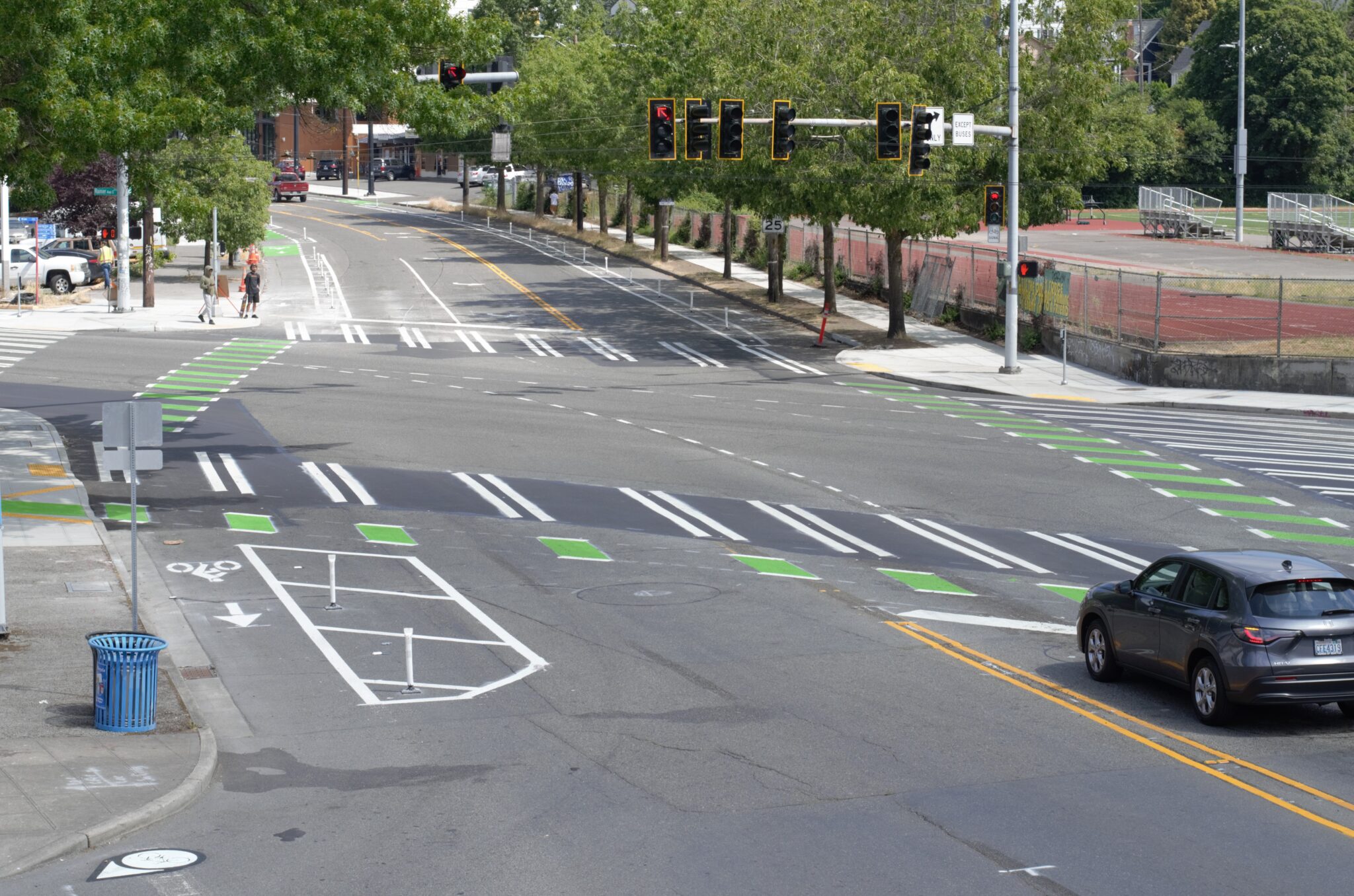 The intersection of MLK Jr Way S and Rainier Ave S features new protected bike lanes, crosswalks, bike signals, and enhanced street markings, designed to improve safety and accessibility. Trees and urban surroundings frame the scene.