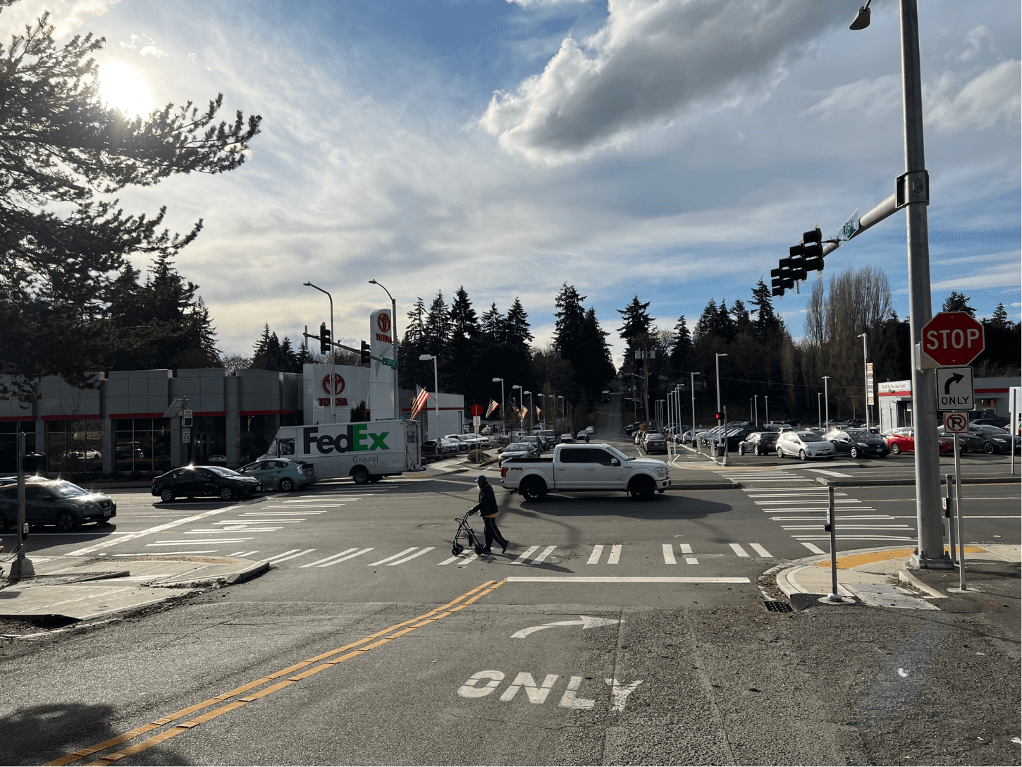 A sunny street intersection with several cars and a person using a walker crossing the crosswalk. A car dealership with parked cars is visible in the background, along with evergreen trees and a partly cloudy sky.