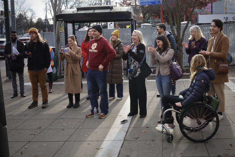 A group of people gathers at a transit stop near Mount Baker Transit Center, smiling and clapping during what appears to be a celebratory event.
