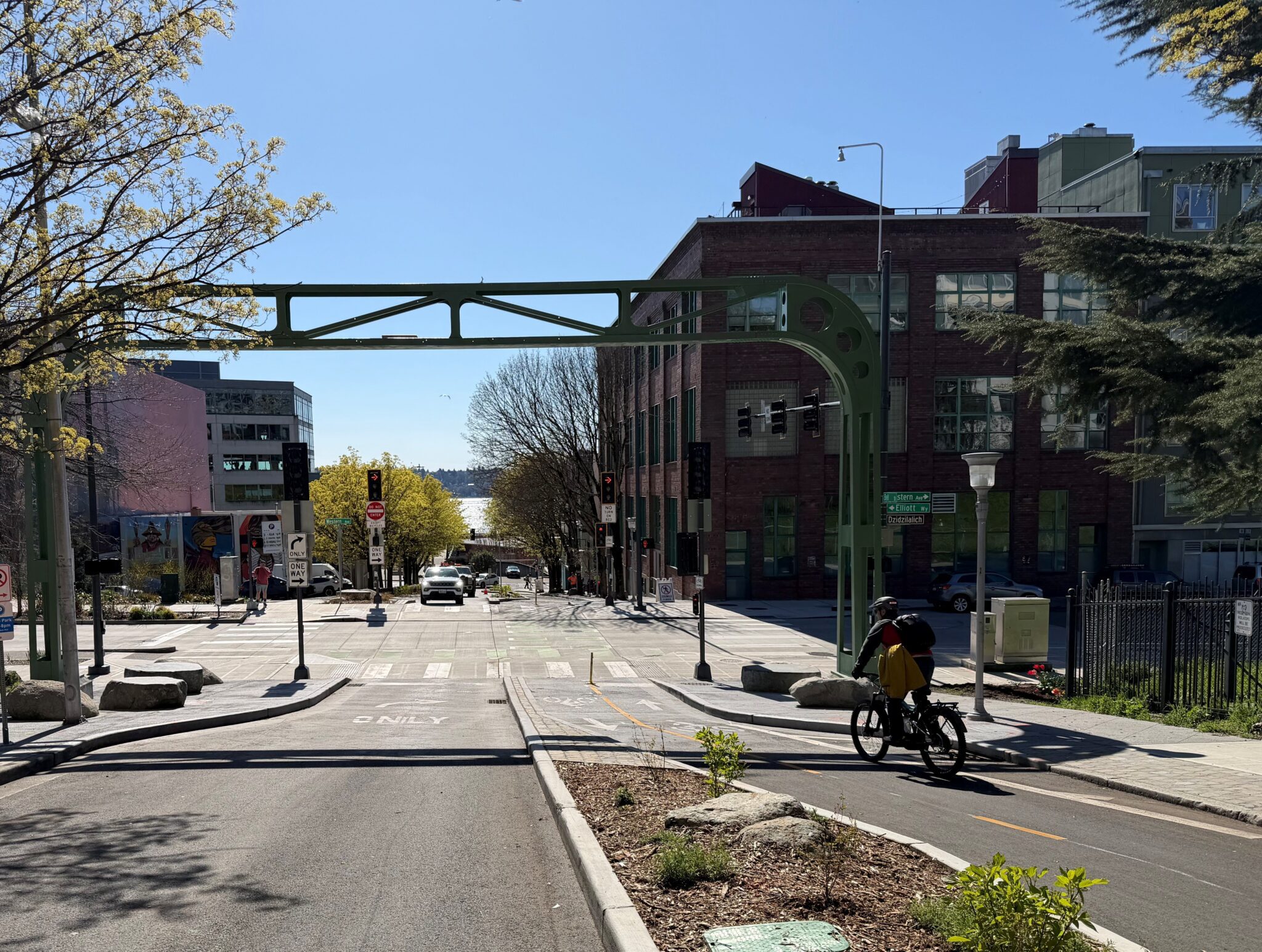 A green, metallic arch stands over a roadway that slopes downward and with a landscaped median in the center. On the right side of the frame, a person rides a bicycle towards an intersection in the center of the frame.